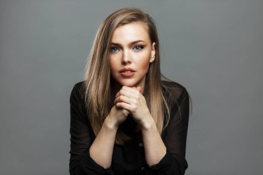 Ciddi genç bir kadın elleri bağlı olarak masada oturuyor. Güzel sarışın. Kariyer ve iş. Gri arkaplan.