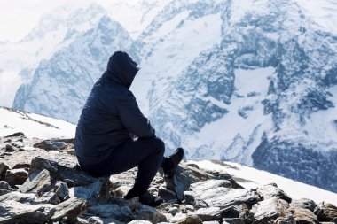 Karlı bir dağın yamacında oturan bir adam. Kapüşonlu mavi ceketli adam. Aktif turizm, yürüyüş ve kış sporları. Güzel manzara.