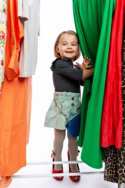 Askıdaki elbiselerin arasında kırmızı ayakkabılı, gülümseyen küçük bir kız. Zevk, mutluluk ve moda. Dikey.