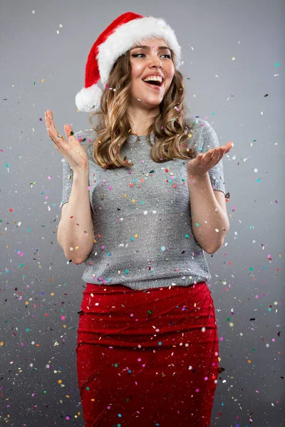 一个戴着圣诞礼帽的漂亮女人在一场五彩纸屑的雨中欢笑着 享受着节日 传统的冬季活动 圣诞节和新年 灰色背景 垂直方向 — 图库照片