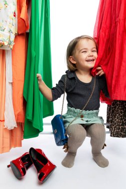 Askıdaki elbiselerin arasında kırmızı ayakkabılı, gülümseyen küçük bir kız. Zevk, mutluluk ve moda. Dikey.