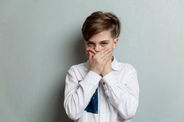 The boy covered his mouth with his hands. Emotional child in a white shirt on a background of a gray wall. Fear and horror.