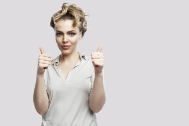 Smiling young woman with thumbs up. Beautiful blonde in a gray shirt. Success and positive. Isolated on a light gray background. Space for text.