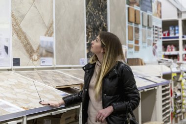 Genç bir kadın bir hırdavatçıda seramik fayanslar seçiyor. Siyah ceketli güzel bir sarışın. İnşaat, tasarım ve onarım.