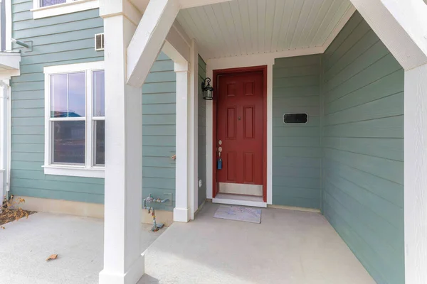 Townhouse porch with lockbox on the red front door. Entrance exterior of a townhouse with greenish gray wood vinyl siding and white trims on the windows and columns.