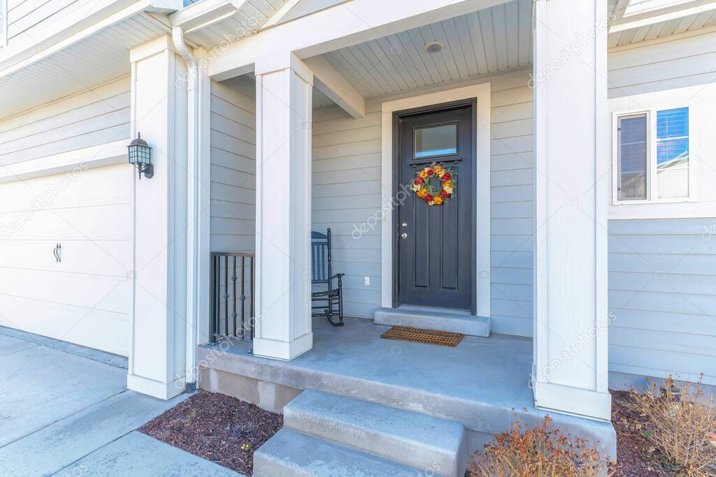 House porch with rocking chair near the black front door with glass panel and wreath. Exterior of a house with light gray vinyl lap sidings and white garage door on the left near the porch.