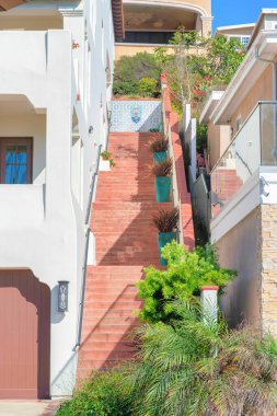 Binanın dışında kırmızı beton merdivenler ve her inişte saksı bitkileri var. Merdivenlerin önünde soldaki beyaz binada tırabzan olan bitkiler var..