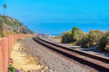Solunda tahta direkler olan San Clemente, California 'daki tren rayları. Sağdaki çalıların yanında demiryolu ve arkada açık mavi gökyüzüne karşı bir yamaç ve rıhtım manzarası..