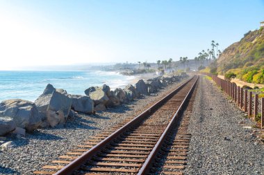 San Clemente, California 'da plaj manzaralı ve yamaçlı tren rayları. Sol tarafta büyük kayalar var ve dağın eteklerinde gökyüzüne bakan palmiye ağaçları var..