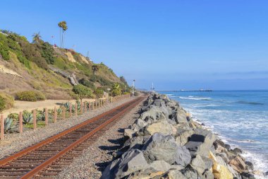 San Clemente, California 'da sahile yakın bir tren yolu. Soldaki dağ yamacına yakın tahta direkli demiryolu ve arkadaki mavi gökyüzüne karşı rıhtımın manzarası..