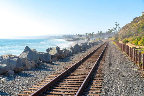 San Clemente, California 'da plaj manzaralı ve yamaçlı tren rayları. Sol tarafta büyük kayalar var ve dağın eteklerinde gökyüzüne bakan palmiye ağaçları var..