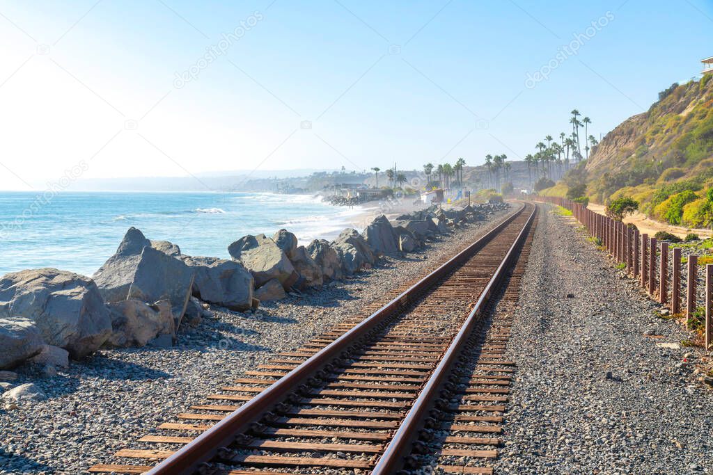 Train track in San Clemente, California with a view of the beach and slope on the side. There are large rocks on the left side and a view of palm trees near the mountain against the sky at the back.