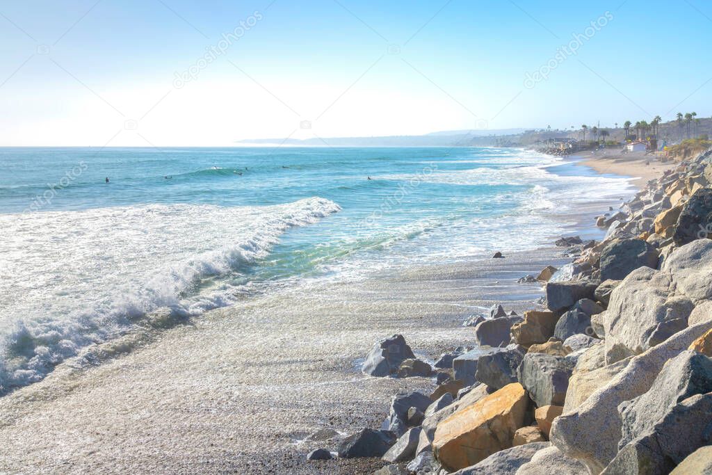 San Clemente beach in California with rocks on the shore. There are people enjoying the waves of the ocean and a view of palm trees and mountain against the sky at the back.