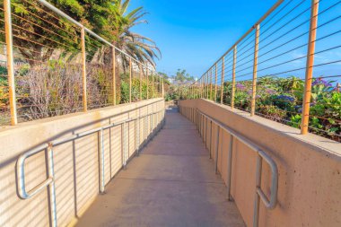 San Clemente, Kaliforniya 'da duvarların arasına giden yol. Duvarların yanında zemine monte edilmiş metal parmaklıklar ve temiz gökyüzü arka planında bitkiler olan beton yol..