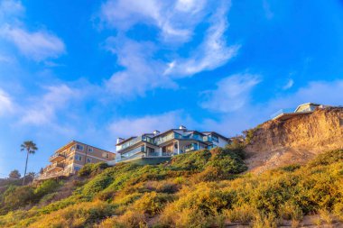 San Clemente, CA 'nın tepesinde yabani bitki ve binaların bulunduğu bir dağ yamacının alçak açılı görüntüsü. Solda gökyüzüne karşı balkonları olan iki apartman var..