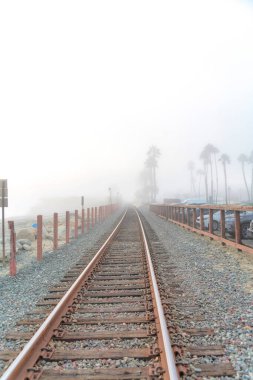 San Clemente, California 'nın sisli sahilinde tren yolu. Solda direkler ve sağda tahta çitler var. Park halindeki araçların yanında..