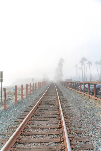San Clemente, California 'nın sisli sahilinde tren yolu. Solda direkler ve sağda tahta çitler var. Park halindeki araçların yanında..