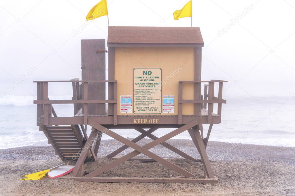Wooden lifeguard tower with two yellow flags on top against the foggy beach of San Clemente, CA. Lifeguard tower exterior with advisory signage on the wall and keep off sign on the beam.