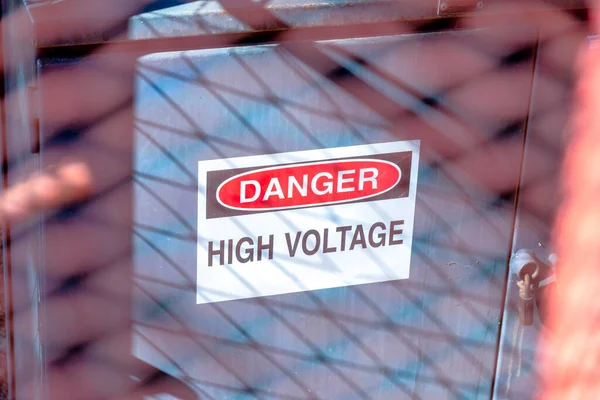 Danger high voltage sign on a gray electrical fuse box at San Jose, California. Warning sign posted at the door with a mesh wire fence shadows.