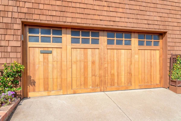 Wooden bi-fold garage doors with glass panel at San Francisco, California. Garage exterior with plants at the side of the concrete driveway and a brown shingle sidings on the wall.