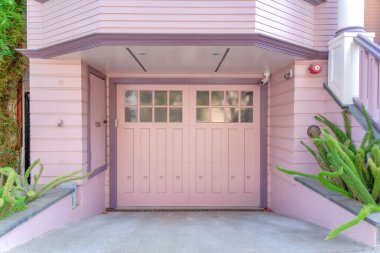 Garajın dış tarafında çift menteşeli kapı ve San Francisco, California 'da pembe tahta kaplama var. Garaj yolunun kenarında cam paneller ve kamerayla kapıya doğru giden bitkiler var..