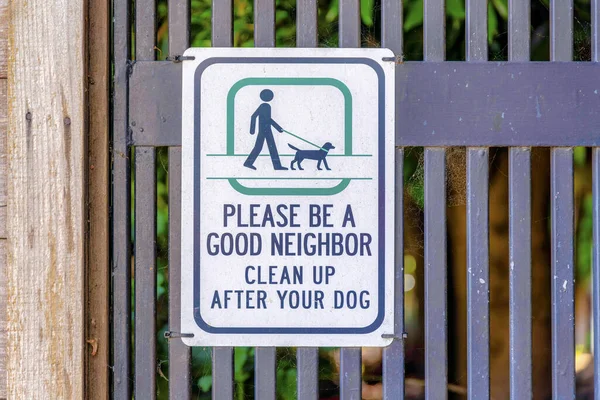 Lütfen iyi bir komşu ol ve köpeğinin arkasını temizle. İşaret, arkasında ağlar ve solunda tahta direkler olan demir parmaklıklara asılmış..