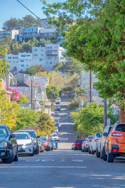 San Francisco, California 'da park etmiş araçlarla dolu eğimli beton bir yol. Yan taraflarında ağaçlar olan dar sokak yolu arkadaki konut binalarının manzarasına karşı..