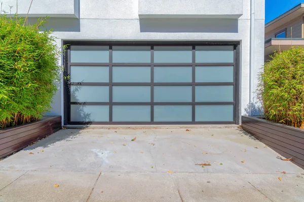 Modern garage door with frosted glass panel and black grids at San Francisco, California. Concrete driveway with small bamboo plants on a wooden black planters on the side.