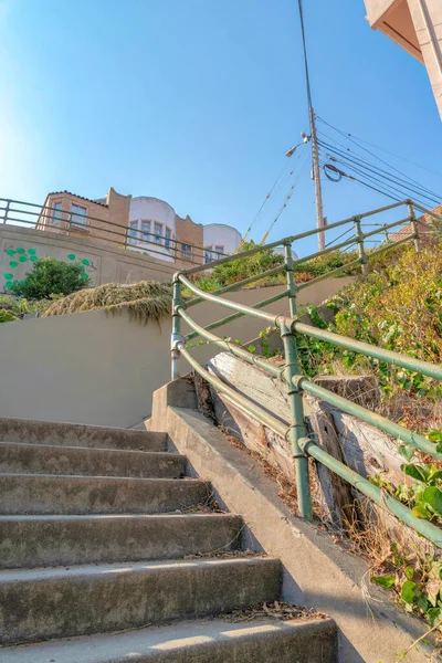 Staircase with concrete steps and painted green metal bar handrails in San Francisco, California. There are grasses on the side of the stairs and a view of electric cables and residential building.