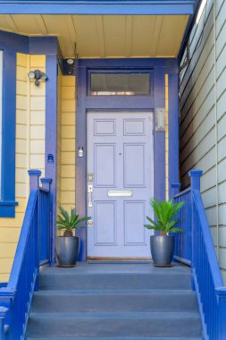 San Francisco, Kaliforniya 'da mavi süslemeli ve sarı ahşap kaplamalı bir evin dışı. Mor kapının önünde iki saksı bitkisi olan verandaya çıkan bir merdiven var..