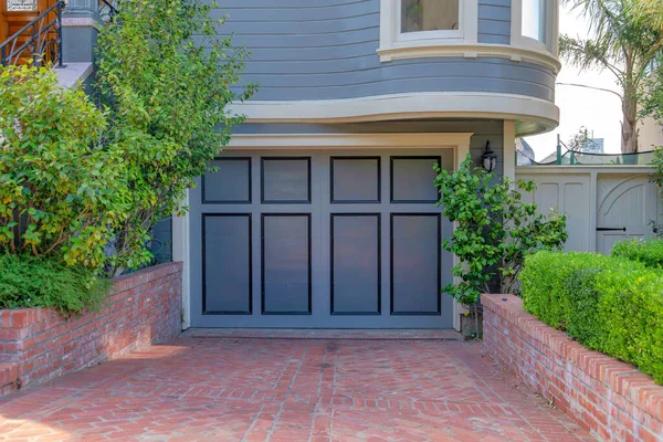Garage exterior with side hinged doors and gray wall sidings at San Francisco, California. Driveway and walls with bricks leading to the garage door with dark gray panels and black trims.