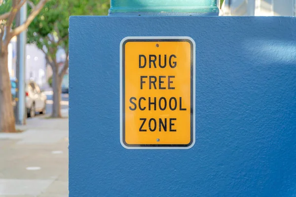 Drug free school zone on a yellow signage at San Francisco, California. Yellow signage on a bluish gray concrete wall with a view of a pavement, trees, and vehicles at the back.