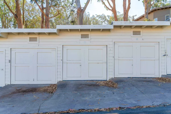 Garage doors on a sloped area in San Francisco, California. Garage exterior with air vents at the top of the doors and white vinyl lap sidings against the trees and sky at the back.