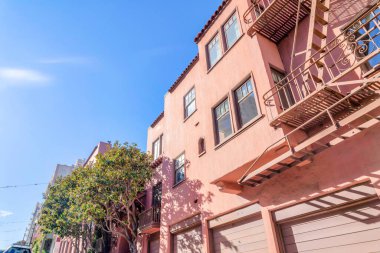 San Francisco, California 'daki açık mavi gökyüzüne karşı alçak açılı apartmanlar. Solda bir bina var. Altında garaj kapıları ve acil durum merdivenleri var..