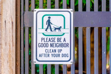 Lütfen iyi bir komşu ol ve köpeğinin arkasını temizle. İşaret, arkasında ağlar ve solunda tahta direkler olan demir parmaklıklara asılmış..