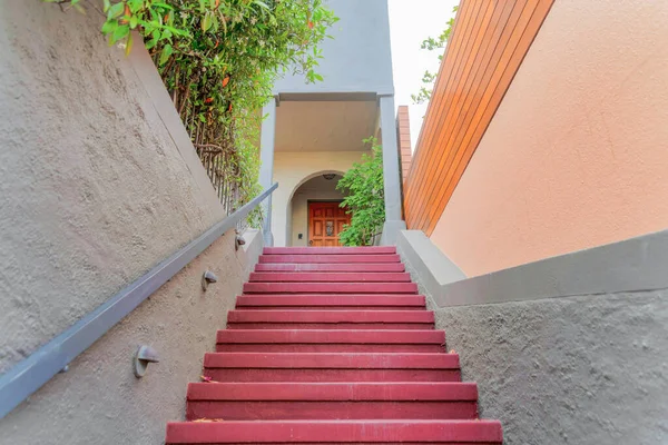 Staircase with red steps and wall-mounted metal railing at San Francisco, California. Stairs in the middle of the walls with plants on the top left side leading to a porch with wooden door.