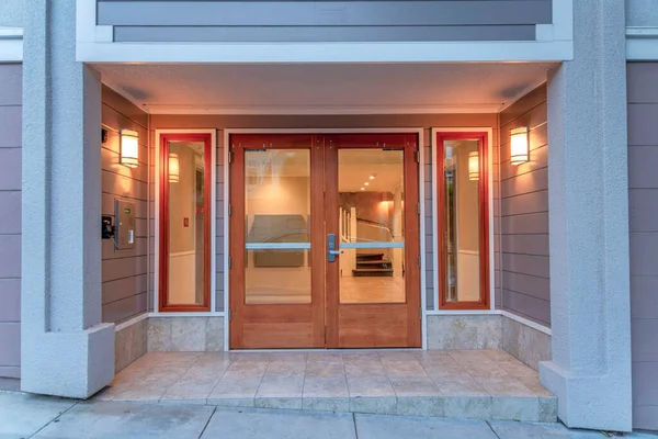 Double door with glass and wood frames of a modern apartment building at San Francisco, CA. Exterior of a building with gray wall sidings and two wall lights at the sides.