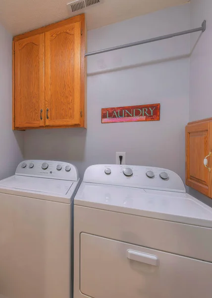 Vertical Laundry room beside the bathroom with large mirror door. Small laundry interior with wall cabinet and metal rod above the two laundry units near the gray wall with signage.