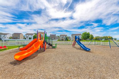 Empty small playground with slides and swings near the houses at Utah Valley. Community playground with white picket fence against the view of houses with trees outdoors.