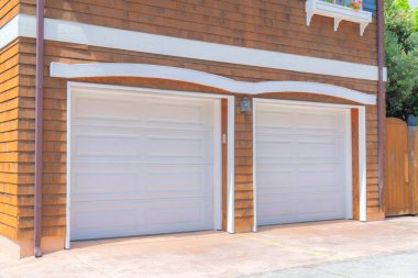 Exterior of an attached garage in La Jolla, San Diego, California. There are two white garage doors with wall lamp in the middle and an exterior of brown shingles sidings.