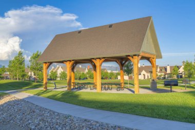 Small pavilion in a large park at Daybreak, South Jordan, Utah. There is a concrete pathways around the pavilion with grill on the side and a view of a lawn and houses against the mountains.