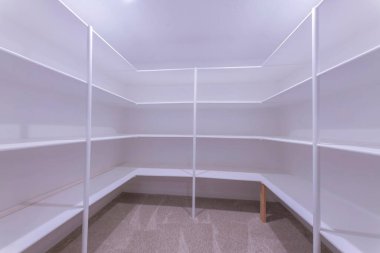 Kitchen pantry with white interior and carpeted flooring. Interior of a pantry with framed shelves on three parts of the wall and stand on the carpet floor.
