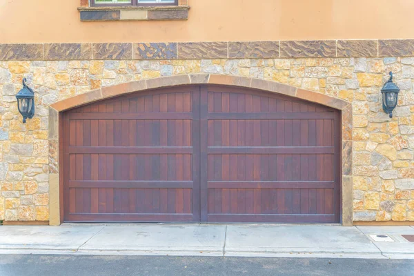 Double wooden garage doors with arched entrance design at Carlsbad, San Diego, California. Garage exterior with stone veneer sidings and two wall lamps at the side of the entrance.