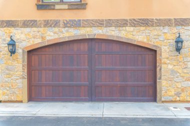 Double wooden garage doors with arched entrance design at Carlsbad, San Diego, California. Garage exterior with stone veneer sidings and two wall lamps at the side of the entrance.