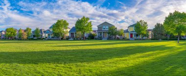 Residential area at Daybreak, Utah with large green fied at the front. There are facades of traditional house structures partially covered by trees against the cloudy sky background.