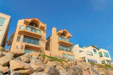 Mediterranean style buildings with window walls at Oceanside, California. Beachfront houses near the seawall with large rocks and handrails at the front.
