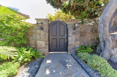 Arched black front door gate in the middle of two wall posts with lamps and doorbell. Residence at La Jolla in California with stone walls and path with landscape at the front.