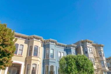 Complex townhouses with bow windows in a low angle view in San Francisco, California. There is a building on the right with emergency stairs and two buildings on the left with beige and blue exterior.
