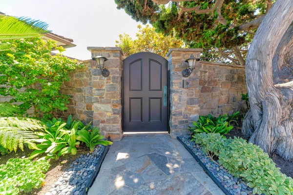 Arched black front door gate in the middle of two wall posts with lamps and doorbell. Residence at La Jolla in California with stone walls and path with landscape at the front.