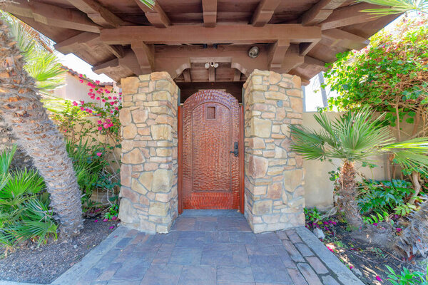 Wooden door gate with cctv on the ceiling at La Jolla, California. Door in the middle of posts with stone sidings and stone tile path with plants and trees on the sides.